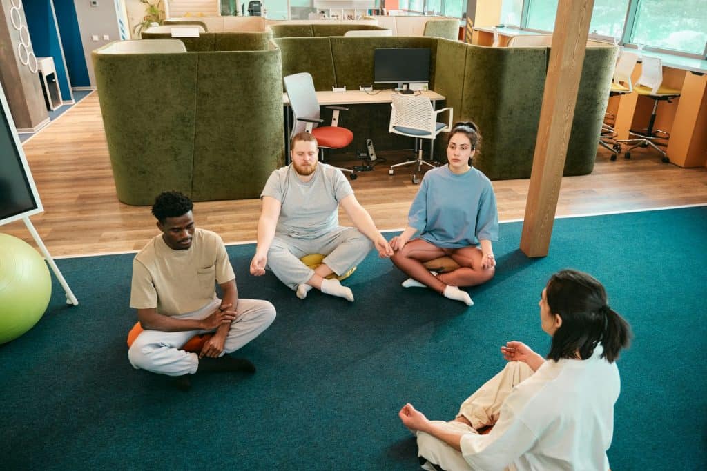 A group of people sitting cross-legged on the floor in a modern office space, participating in a meditation session with a guide.