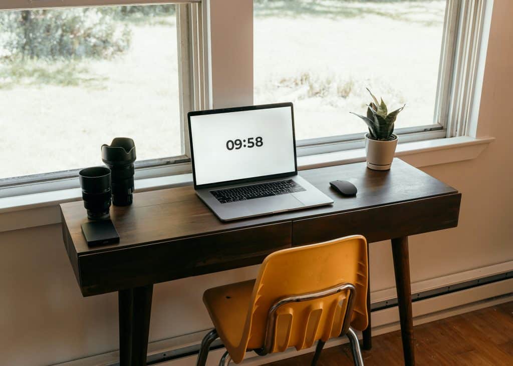 A minimalist workspace view with a wooden desk, laptop displaying the time "09:58," camera lenses, and a potted plant, situated by a window with natural light.