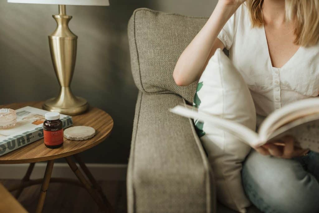 A woman sitting on a couch, reading a book, with a wooden side table holding a bottle of probiotics, some magazines, and a lamp, suggesting a cozy reading nook.