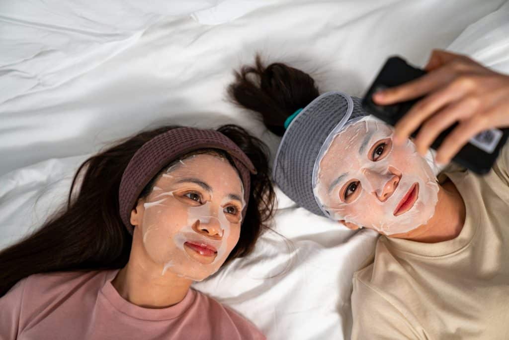 Two women lying on a bed with sheet masks on their faces, one taking a selfie with a smartphone while the other looks on, enjoying a self-care moment.