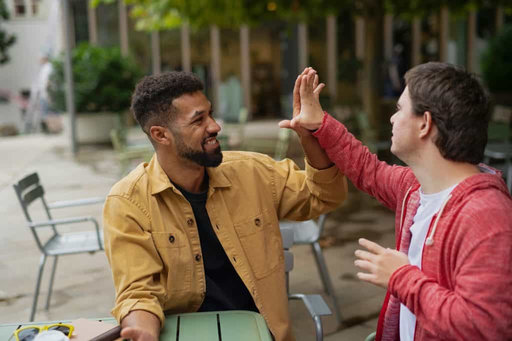 Two men sitting outdoors at a table, giving each other a high-five and smiling, with a relaxed and friendly atmosphere.