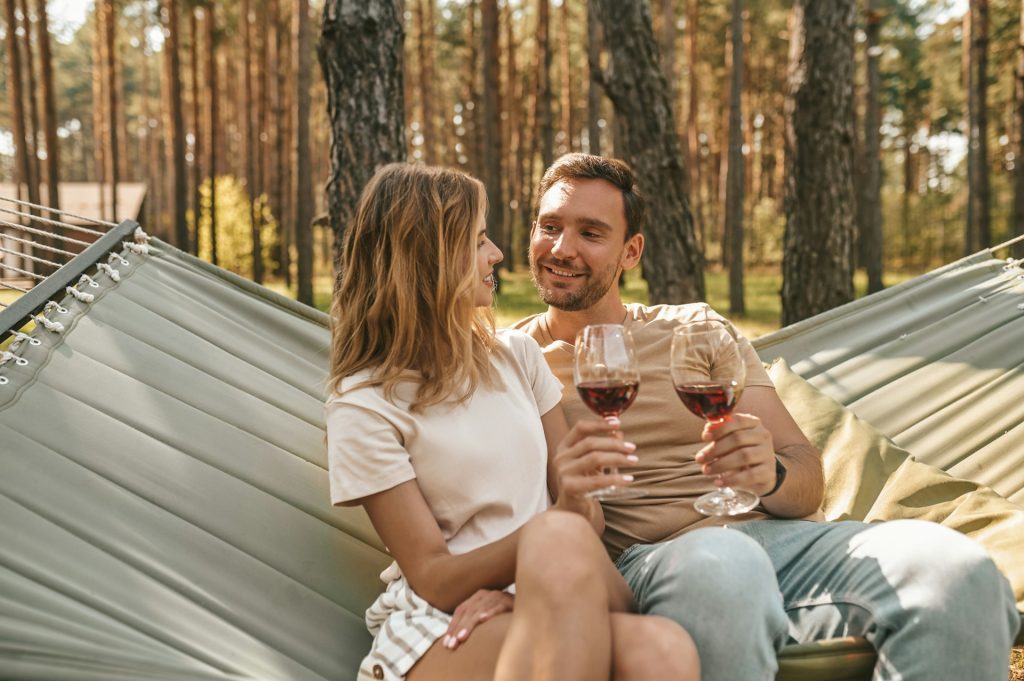 A couple sitting on a hammock in a forest, smiling at each other and clinking glasses of red wine.