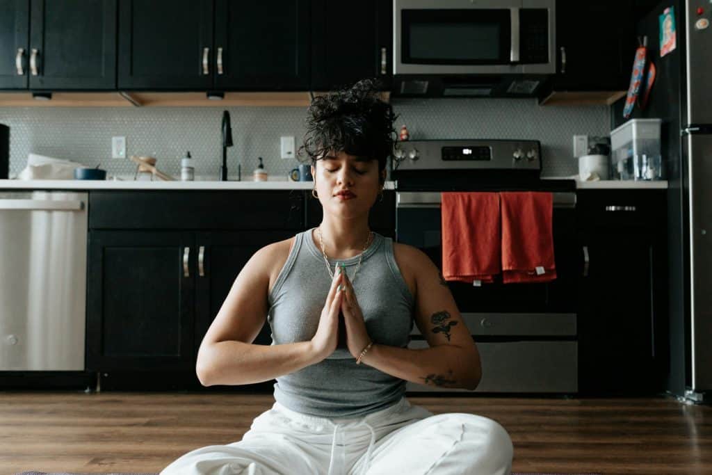 A woman sitting cross-legged in a kitchen, meditating with her hands in a prayer position and eyes closed, creating a peaceful and centered moment.