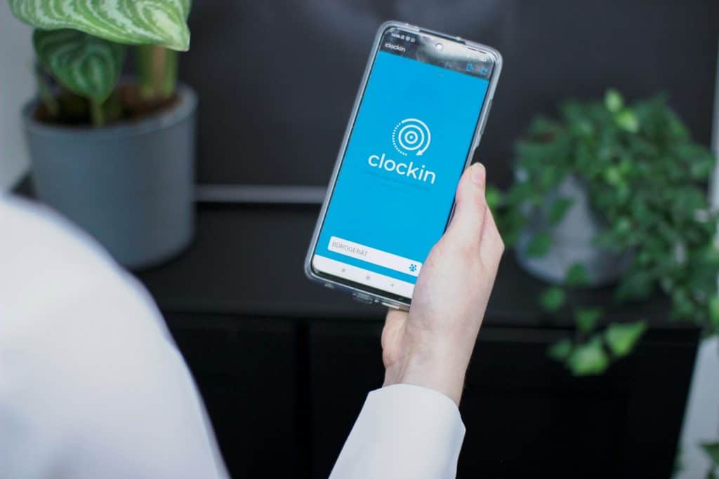 A person holding a smartphone displaying a clock-in app with a blue screen, with plants in the background, suggesting a modern work environment.