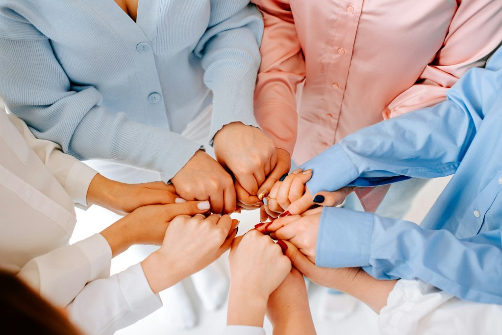 A close-up of multiple hands with interlocking fingers in the center, forming a circle. The hands belong to people wearing colorful button-down shirts.