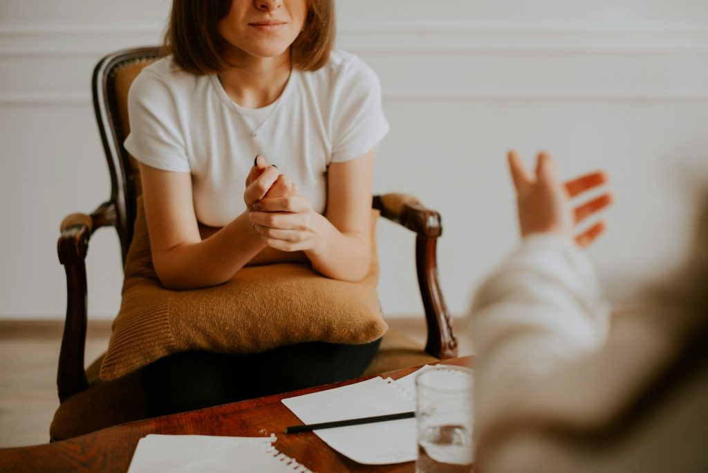 A person sitting in a chair, holding their hands together and talking to someone whose hand is gesturing in the frame; there are papers and a glass of water on the table between them.