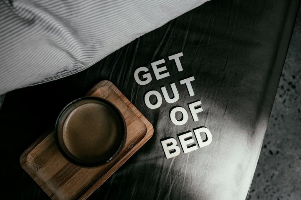 A close-up of the words "GET OUT OF BED" written in letters on a dark grey surface next to a cup of coffee on a wooden tray, promoting motivation.