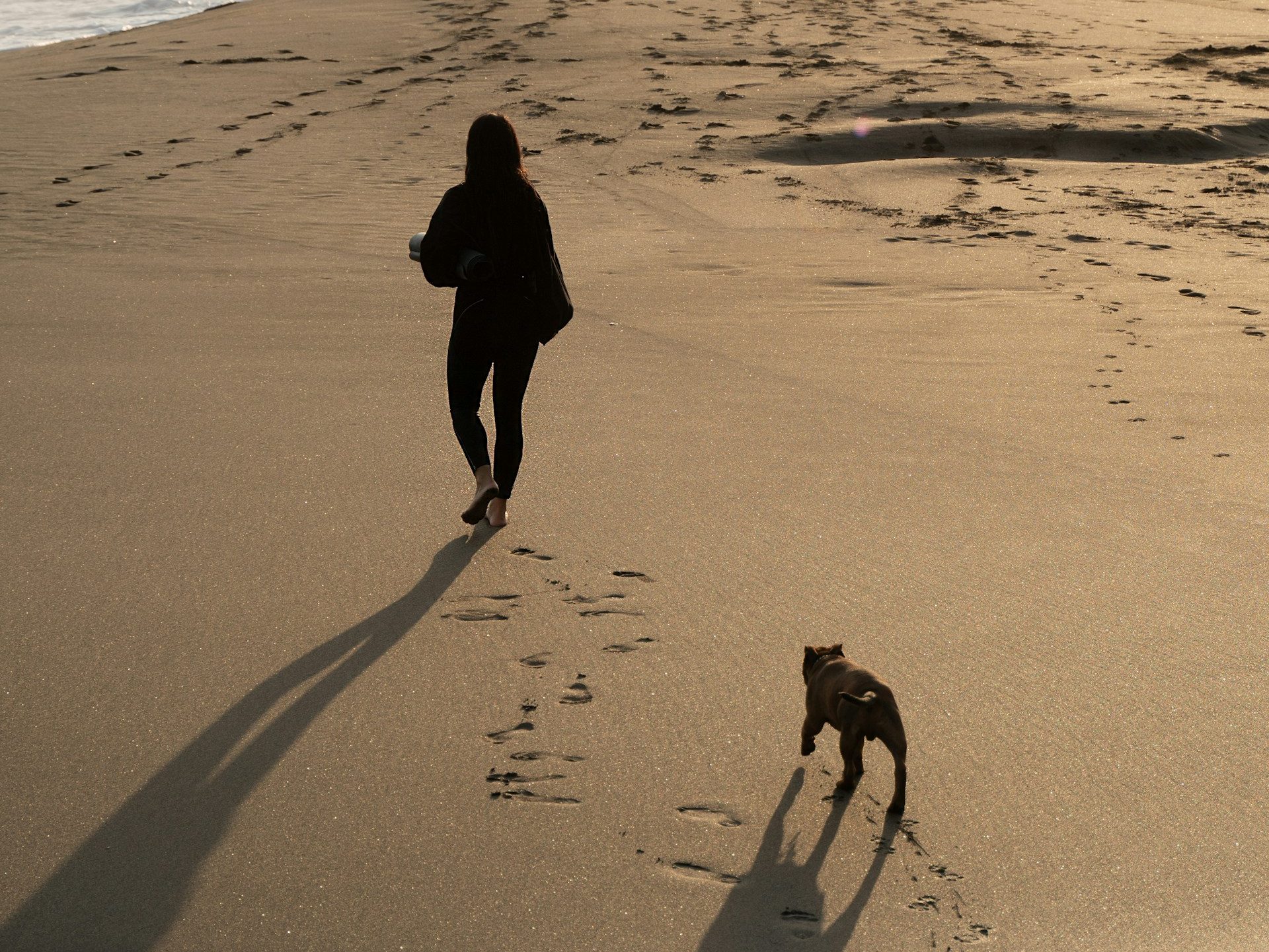 A person walking barefoot on a sandy beach at sunset, accompanied by a dog. Footprints are visible in the sand, with waves gently washing up near the shore.