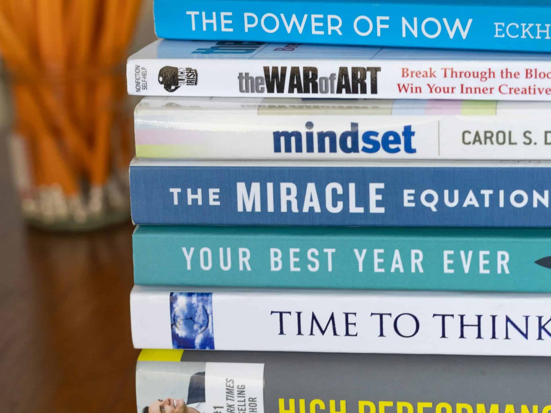 A stack of self-help and motivational books with titles like "The Power of Now," "The War of Art," and "Mindset," with a jar of pencils in the background.